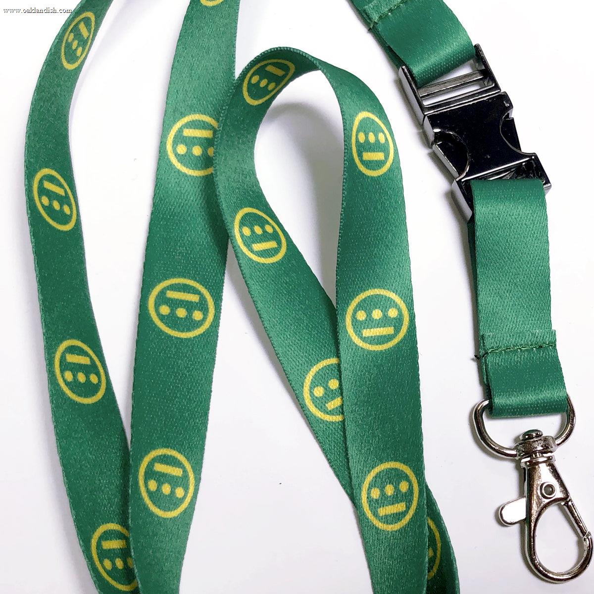 The Hiero VIP Lanyard by Hieroglyphics is a green lanyard adorned with a repeating pattern of yellow, neutral-faced emoji icons that resemble modern hieroglyphics. It features a black plastic quick-release buckle and a metal swivel hook clasp, making it perfect for attaching to your key chain, all set against a white background.