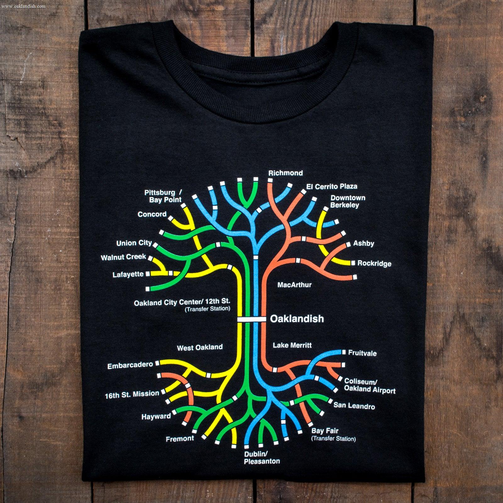 The Oaklandish BART Tree Tee, made from 100% black cotton, is displayed on a wooden surface. This classic fit t-shirt features a colorful, stylized BART map of the San Francisco Bay Area with stations like Civic Center, 12th St., and Concord. The design showcases lines in red, yellow, blue, green, and orange.