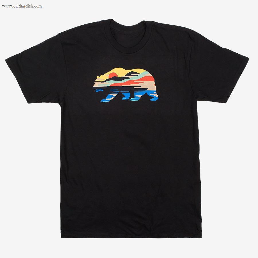 The Sunset Bear Full T-shirt from Never Elsewhere is a black, 100% cotton shirt featuring a stylized design of a California grizzly silhouette filled with a colorful landscape. Inside the bear shape, a red sun, mountains, trees, and blue water are depicted in a horizontal stripe pattern. This T-shirt has short sleeves and a round neck.