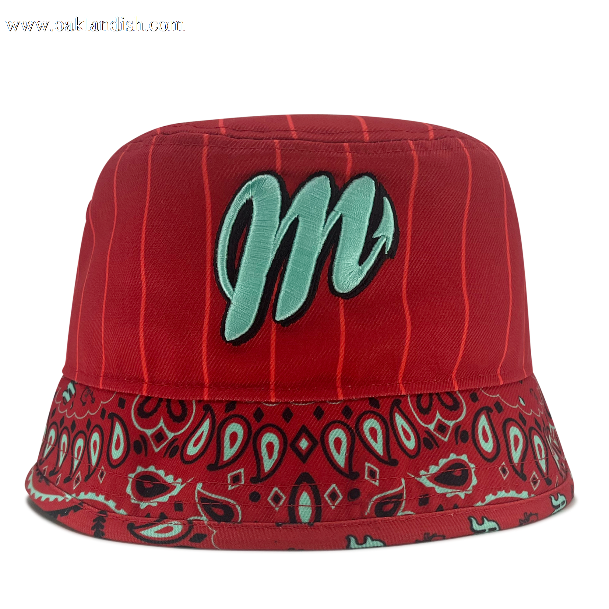 The New Era Diablos Paisley Bucket by Other is a red polyester bucket hat featuring vertical pinstripes, a wide brim with a red, black, and teal paisley bandana pattern, and a large light teal “m” embroidered on the front. The background is plain white.