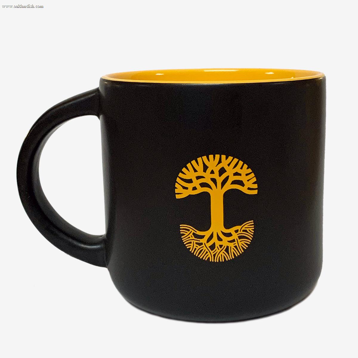 The Sunny Side Mug by Oaklandish is a black ceramic bay mug featuring a glossy yellow interior. Adorning the side is a yellow tree design with branches and roots forming a circular emblem. This stoneware mug boasts a wide handle and a smooth, glossy finish.