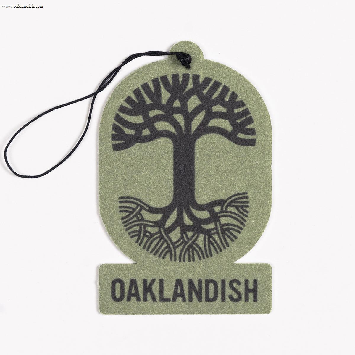 The Oaklandish Air Freshener, from the renowned brand Oaklandish, is a green air freshener featuring a black string at the top for hanging. It showcases a black, stylized tree with spreading roots and branches in the center, enclosed within an oval shape. Beneath the image is "OAKLANDISH" displayed in bold black letters. This air freshener is ideal for those who appreciate unique accessories and delightful scents.