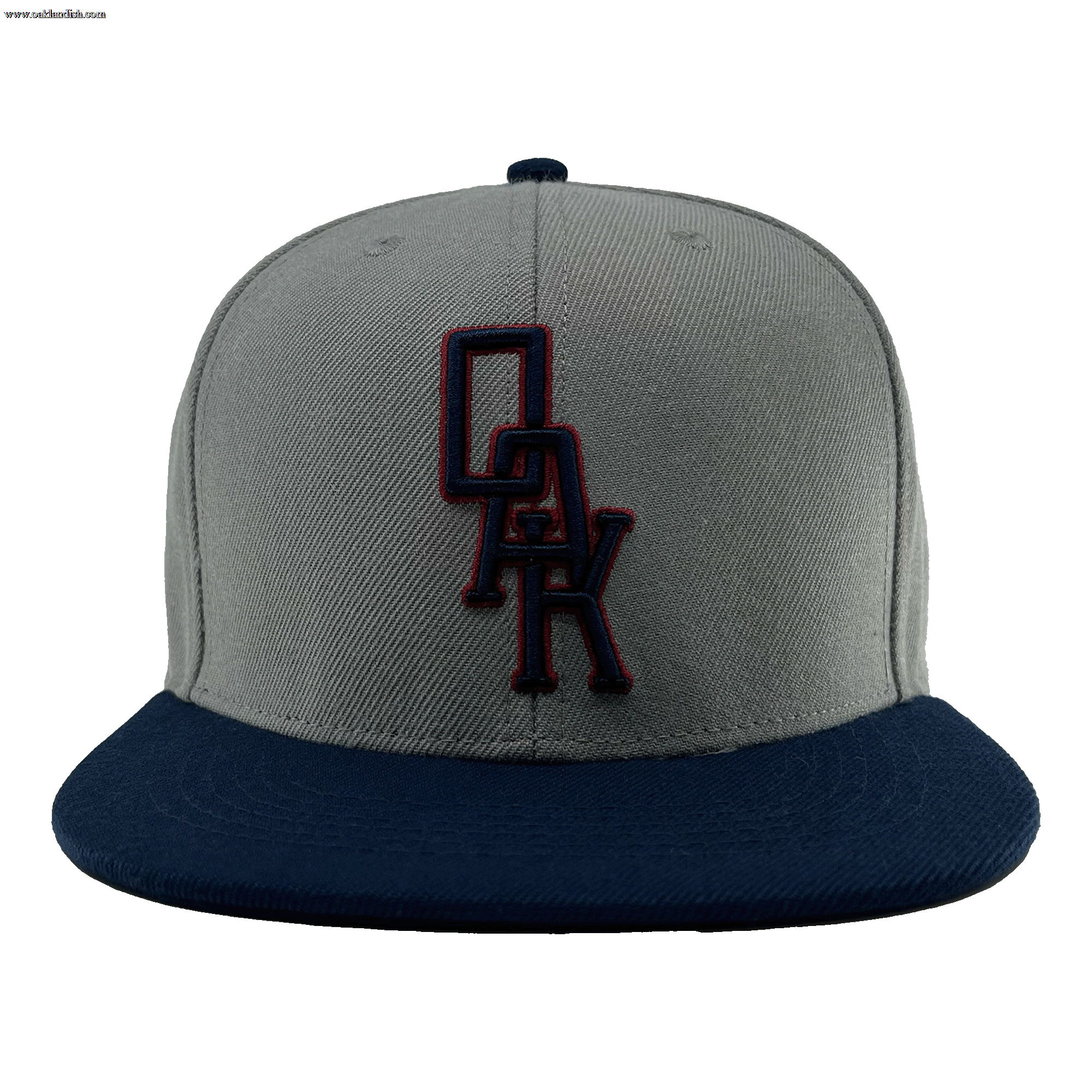 A close-up image of the OAK Monogram Snapback by Oaklandish, showcasing its grey body and navy blue brim. Embroidered in navy blue and outlined in red on the front panel is the distinctive "OAK" logo, evoking Oaklandish style. The cap is centered against a plain green background.