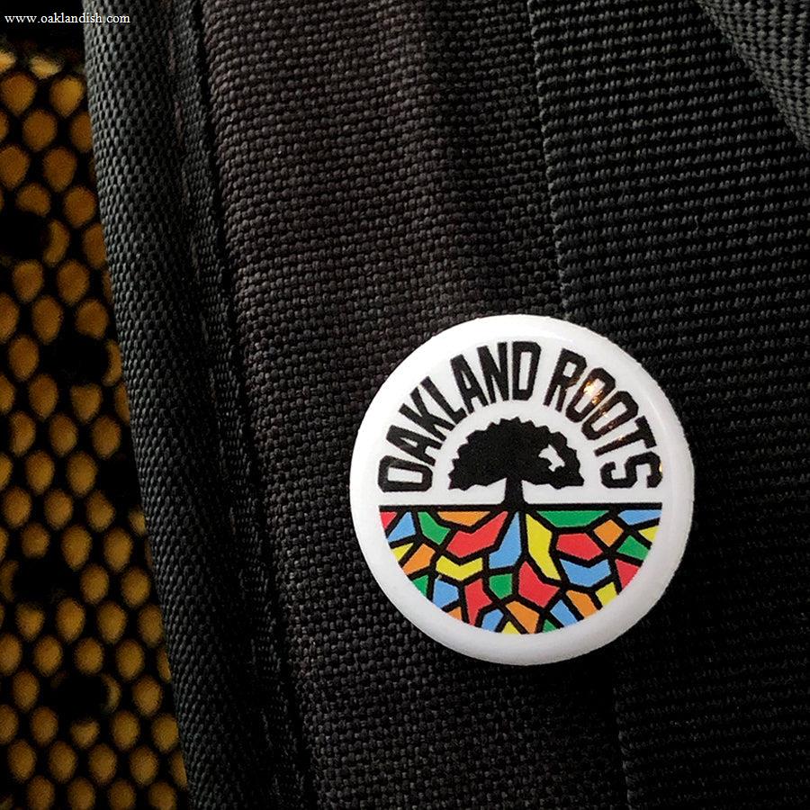 The Oakland Roots SC Pin, a circular badge, is attached to a black woven fabric and proudly displays the "OAKLAND ROOTS" logo. This logo symbolizes a sports club dedicated to social good, showcasing a stylized black tree with roots expanding into a vibrant mosaic pattern at the base. The words "OAKLAND ROOTS" are boldly written in uppercase letters around the top edge.
