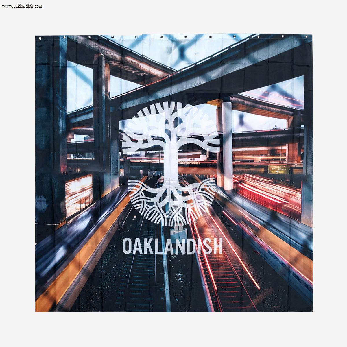 Image showcases the "Overpass Shower Curtain 72 X 72" by Oaklandish. The design features a stylized white tree logo with intertwined roots and branches, overlaid with the word "OAKLANDISH" in white text. The background, reminiscent of a polyester shower curtain, depicts a blurred urban scene with multi-level highways and streaks of light from moving vehicles, evoking a sense of motion and urban energy.