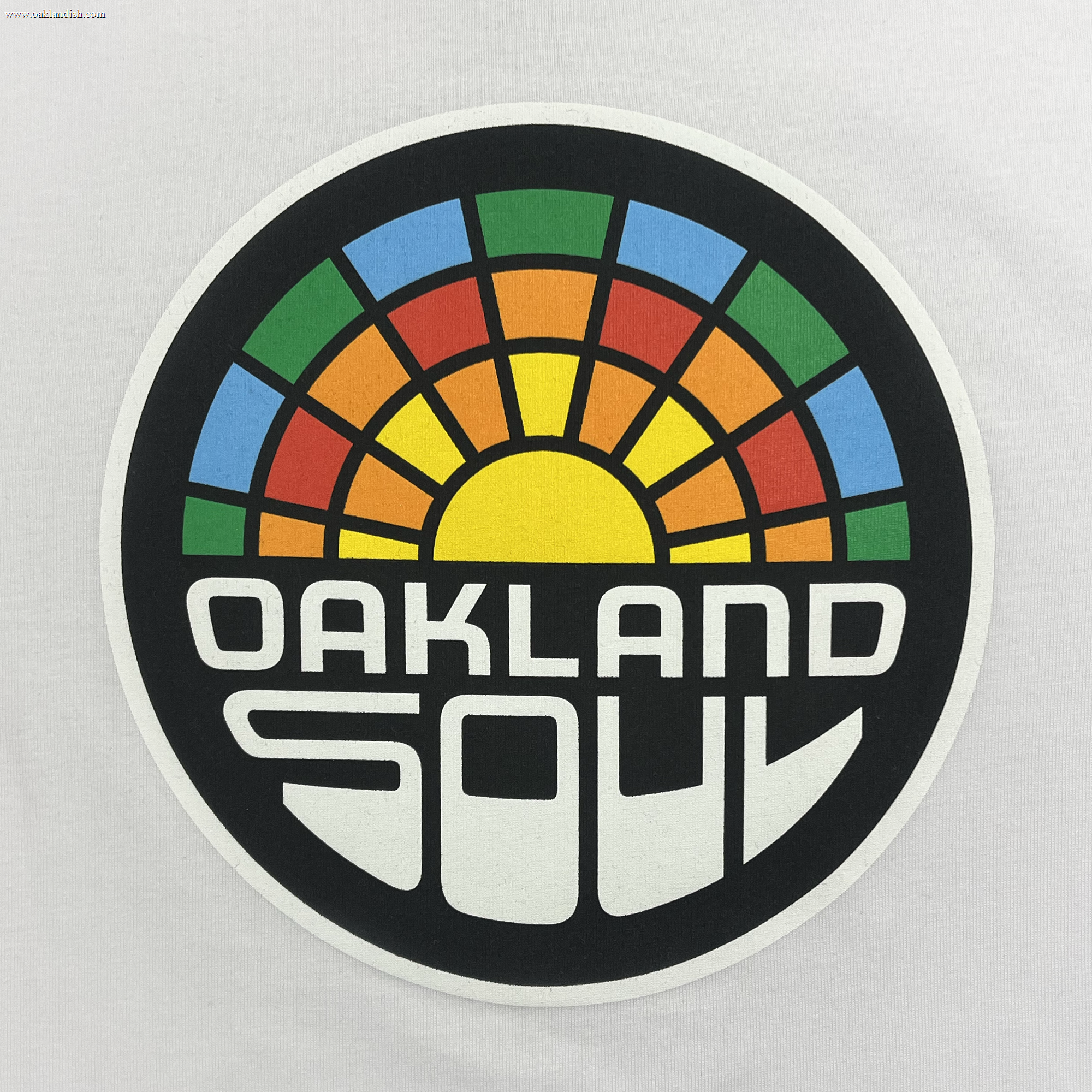 The Toddler Oakland Soul Logo Tee by Oakland Soul features a circular logo on a white background with a stylized, colorful sunrise and a rainbow arch in red, orange, yellow, green, and blue in the upper half. The words "OAKLAND SOUL" are displayed in bold, geometric white letters on the lower half against a black background, representing their USL W-League women's soccer team.