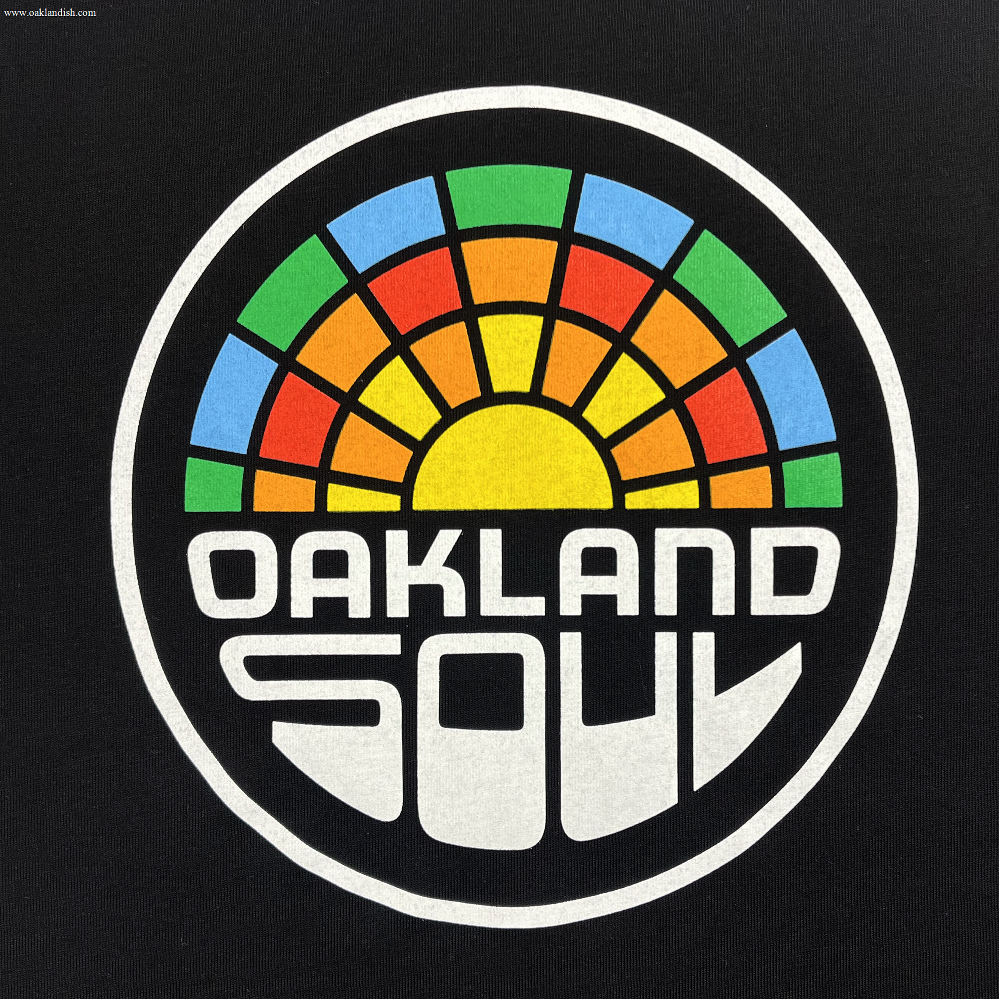 The Infant Oakland Soul Logo One-Piece by Oakland Soul showcases a bold design featuring the words "OAKLAND SOUL" in white, stylized letters at the bottom of a circular logo. Above this text is a vibrant semi-circle resembling a sunset or sun rays with segments in blue, green, yellow, orange, and red. This emblem proudly represents the Oakland Soul women's soccer team in the USL W-League against a sleek black background.