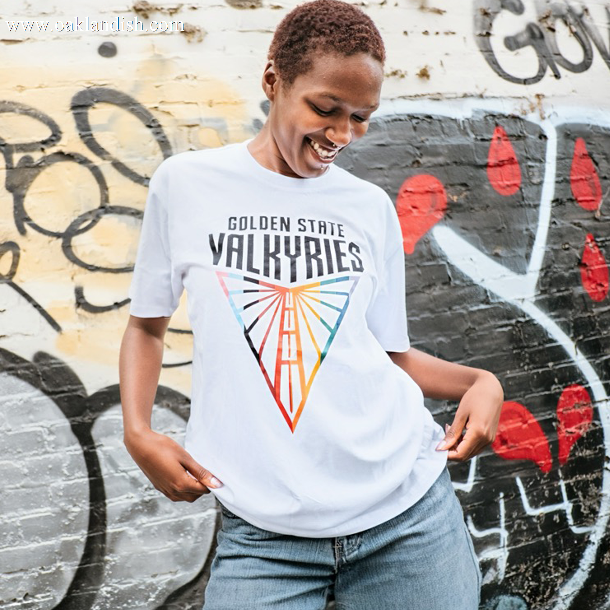 A smiling person with short hair wears the GSV Logo Love Tee by GSV, featuring a vibrant "Golden State Valkyries" geometric design. Made from 100% cotton, they stand before graffiti, holding the hem and looking down in a joyful pose.