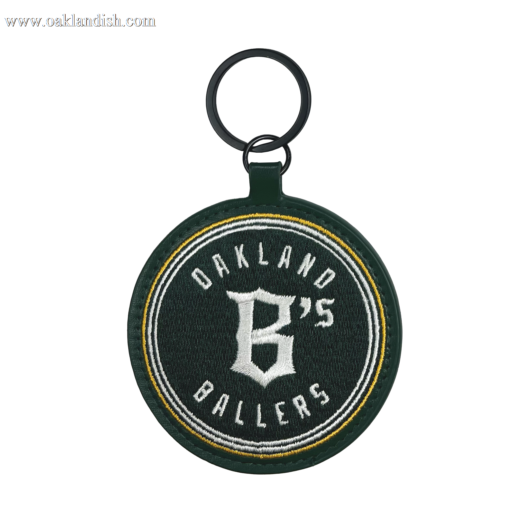 The Oakland Ballers Keychain features a round, dark green fabric embroidered with "Oakland Ballers" in white and a large "B's" at the center. It includes a black ring at the top for attaching keys and has a green border surrounding the entire design. The text is highlighted with yellow and white accent lines, celebrating this pioneering professional baseball team.