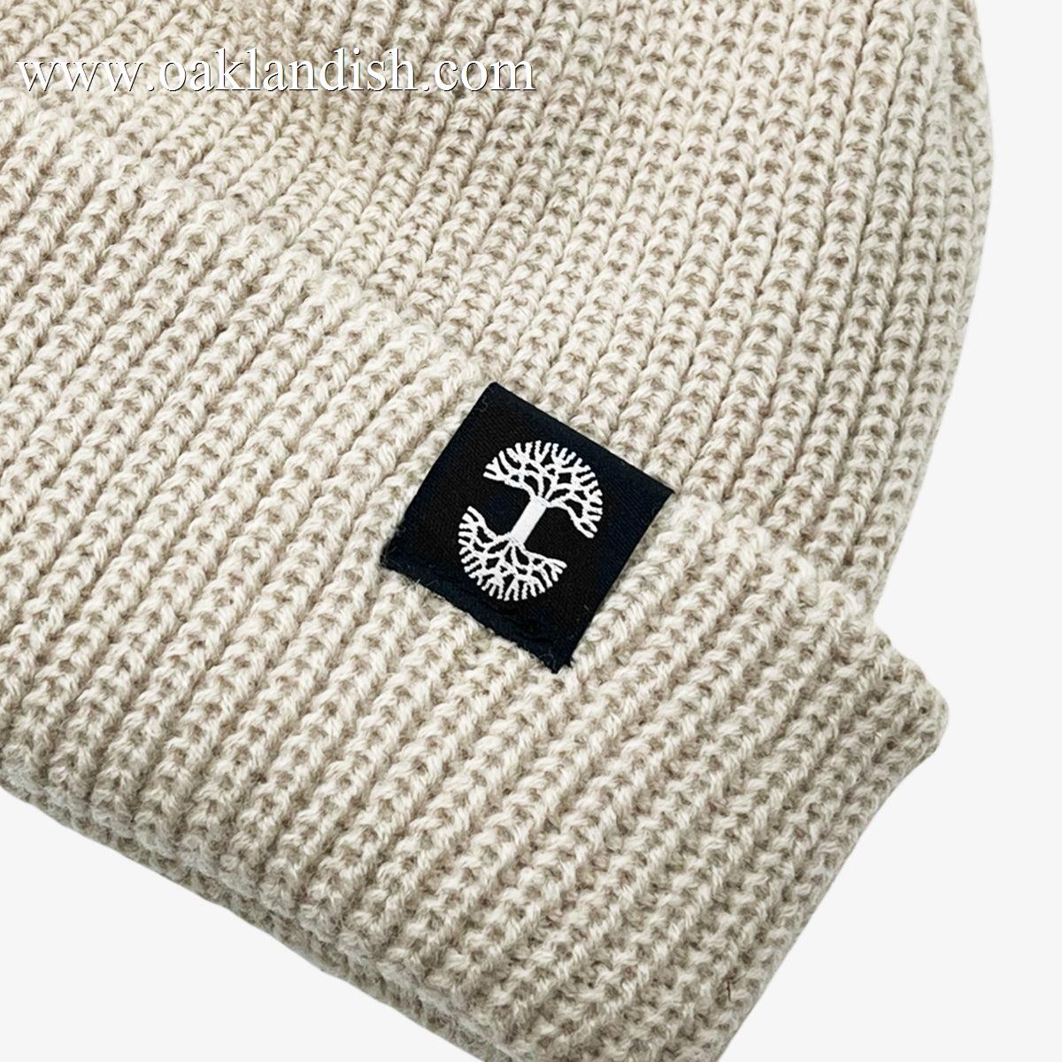 The Ramos Beanie by Oaklandish is a beige knitted hat with a folded brim, featuring a black rectangular patch on the front. The patch displays a white tree emblem with branches forming a circular design around the trunk. This woven acrylic headwear boasts a ribbed texture, giving it a cozy and warm appearance.