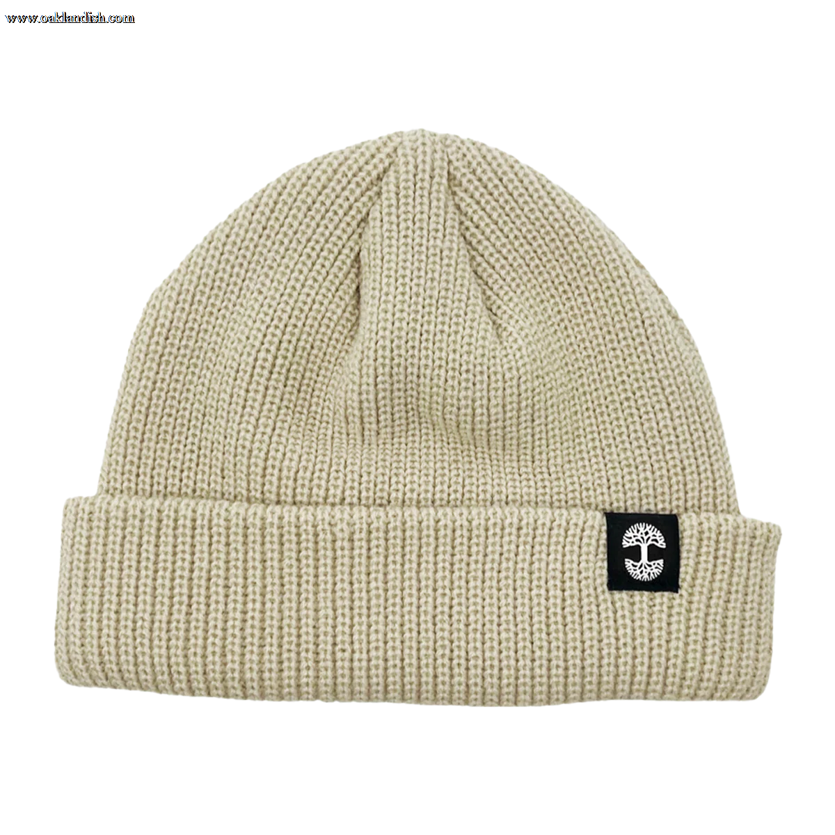 The Ramos Beanie by Oaklandish is depicted, showcasing headwear at its finest with its beige knit and folded brim. The brim features a distinctive black rectangular patch adorned with a white circular cross logo. Made from woven acrylic, the textured material likely provides warmth and comfort. The beanie boasts a simple, classic design that exudes timeless style.