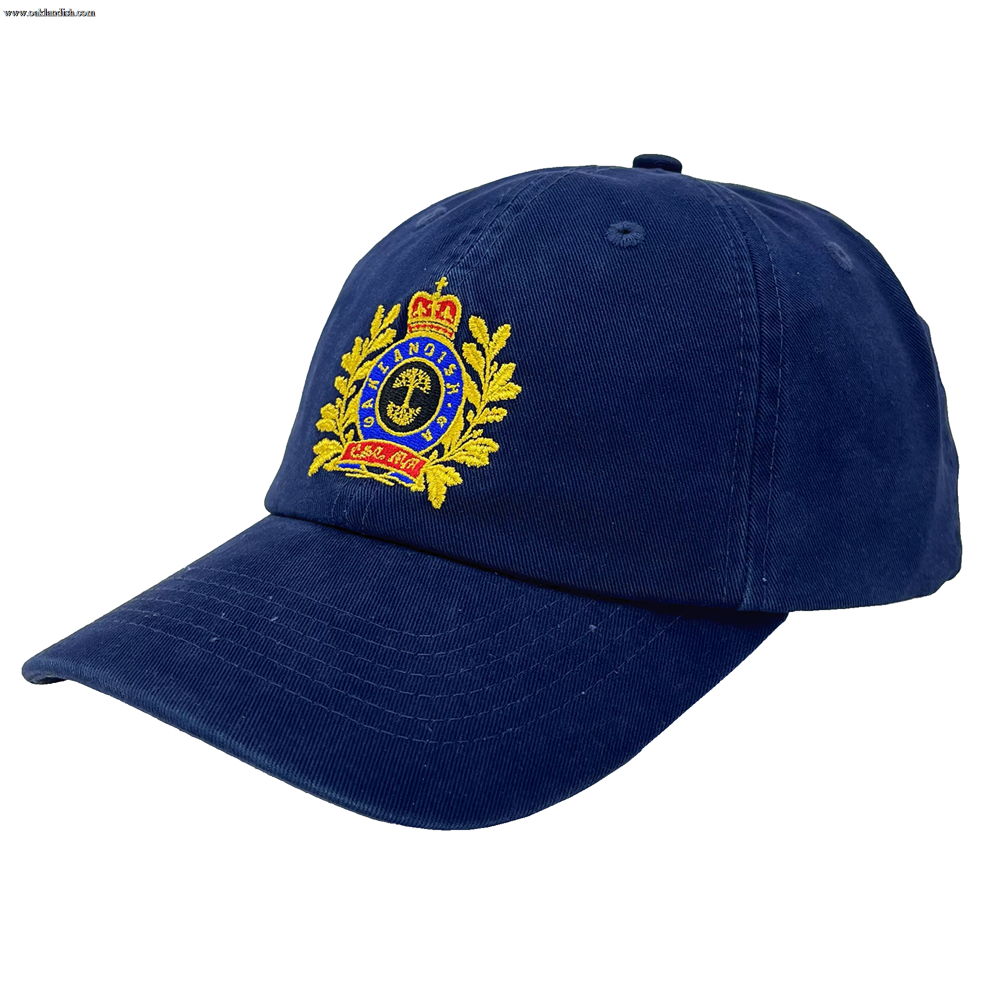 The Regal Crest Dad Hat by Oaklandish is a navy blue, 100% cotton hat with a detailed embroidered emblem on the front. The emblem features a golden crown at the top, a circular design with text below, and laurel leaves on either side. The visor is slightly curved and there’s an adjustable strap at the back for a perfect fit.