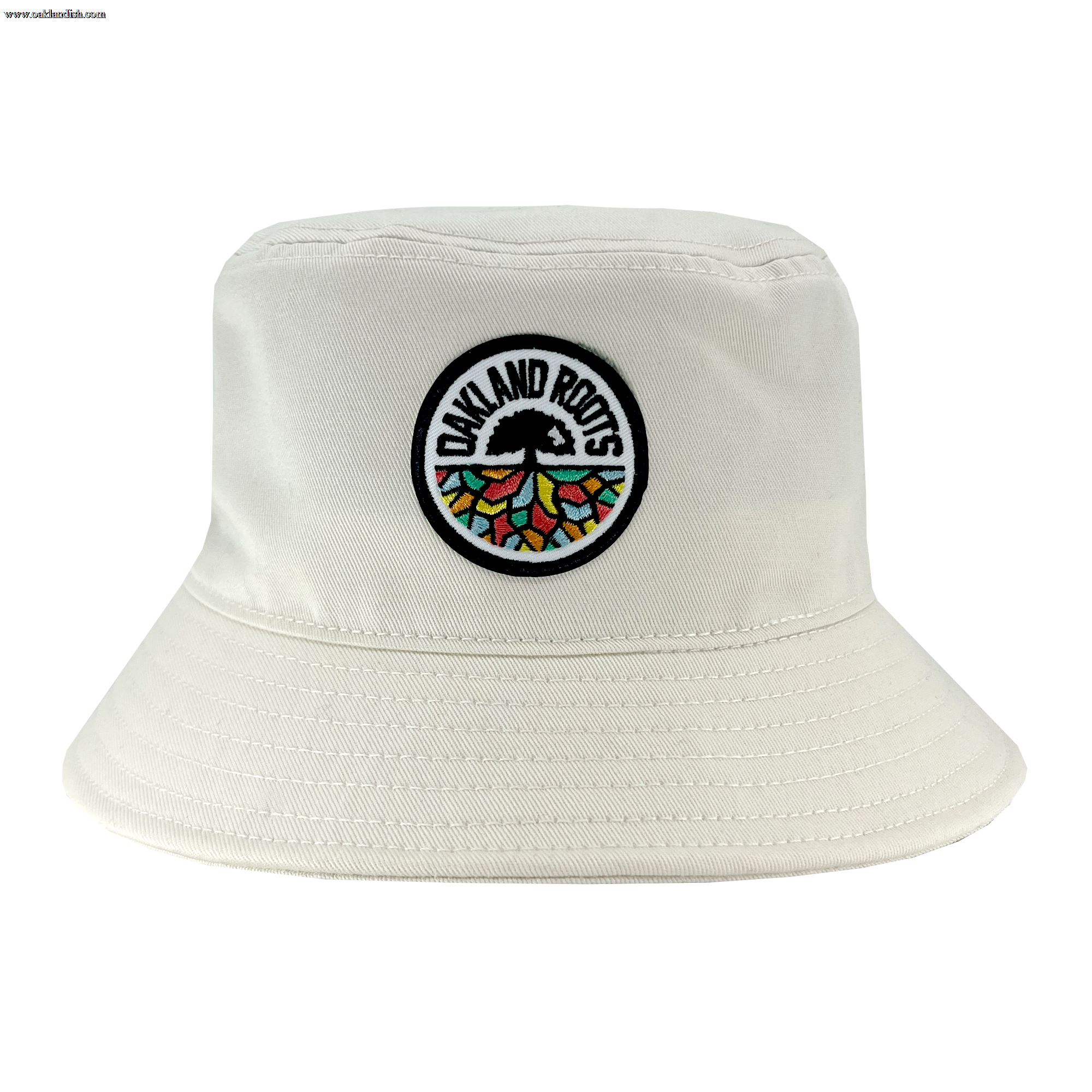 The Oakland Roots SC Bucket Hat, by Oakland Roots SC, showcases a light-colored design with an embroidered circular patch on the front. The patch features a tree with colorful segments and roots, encircled by the text "Oakland Roots," celebrating the local sports club. The hat also includes detailed stitching along the brim and crown.