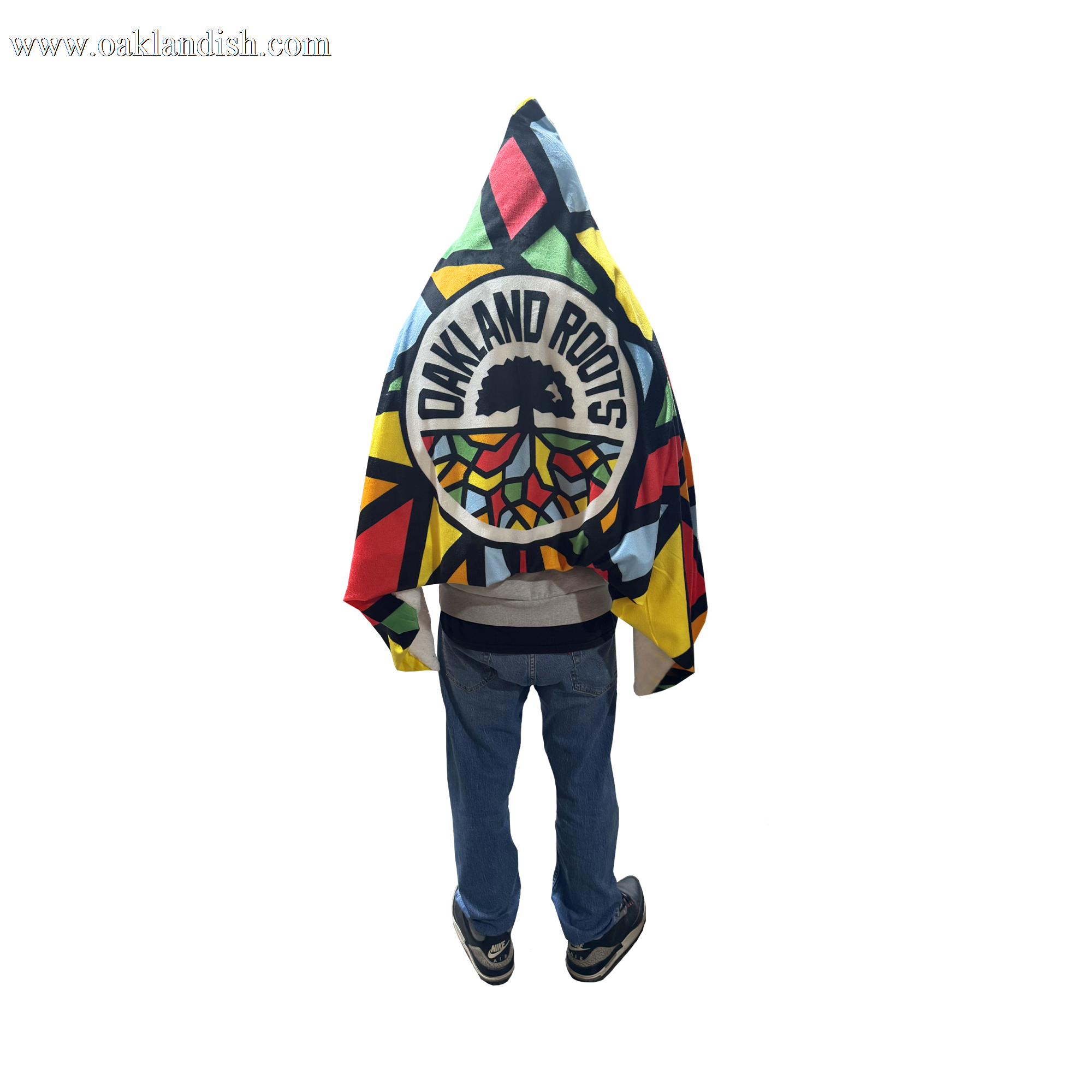 A person stands with their back turned, wrapped in the Oakland Roots SC Sherpa Blanket featuring a bold tree logo and geometric patterns. The cozy blanket, by Oakland Roots SC, pops against their hoodie and jeans on a transparent background.