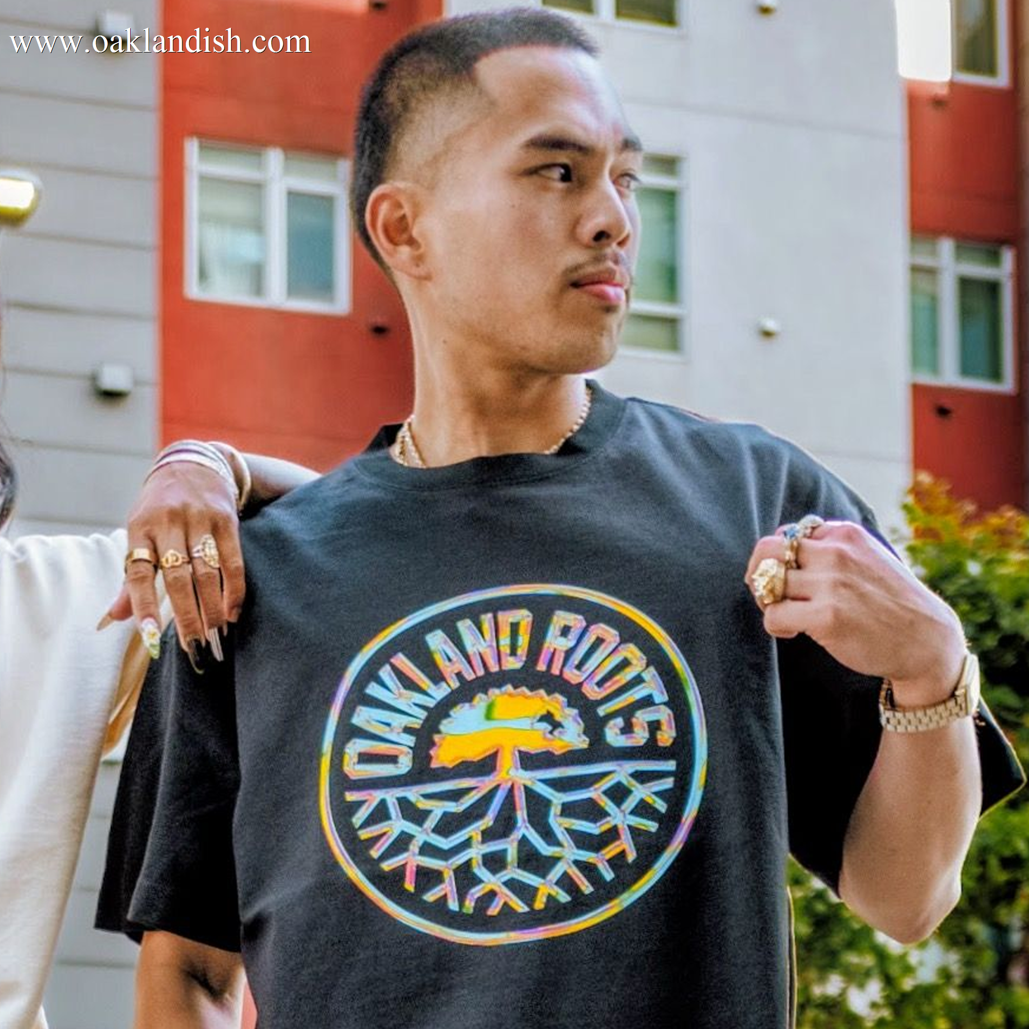 A person stands outdoors by a modern building, looking to the side. They're wearing the Oakland Roots SC Iridescent Tee and pointing to its colorful tree logo. Multiple rings decorate their fingers; another arm is draped over their shoulder.