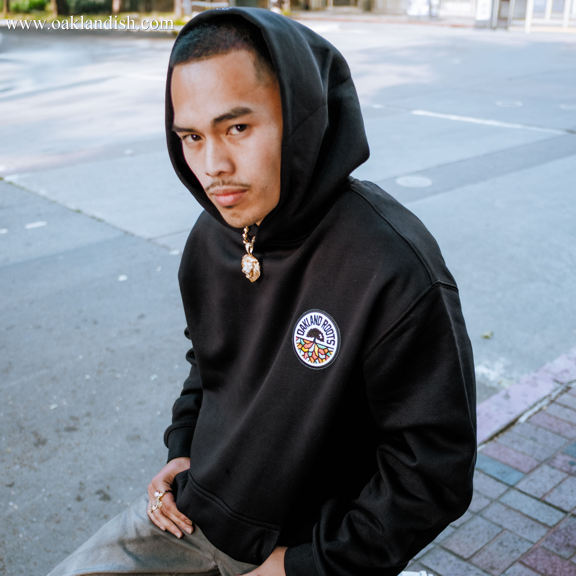 A young man with a shaved head and mustache wears an Oakland Roots SC Logo Premium Hoodie by Oakland Roots SC. Sitting on a city sidewalk, he looks at the camera with rings, a pendant necklace, and a serious expression.