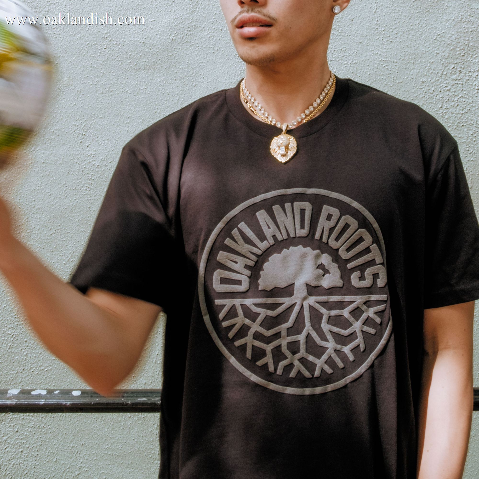 Wearing the Oakland Roots SC Blackout Logo Tee by Oakland Roots SC, a person stands against a light wall. They spin a blurred soccer ball in one hand, sport a gold chain necklace, and their face is partially out of frame.