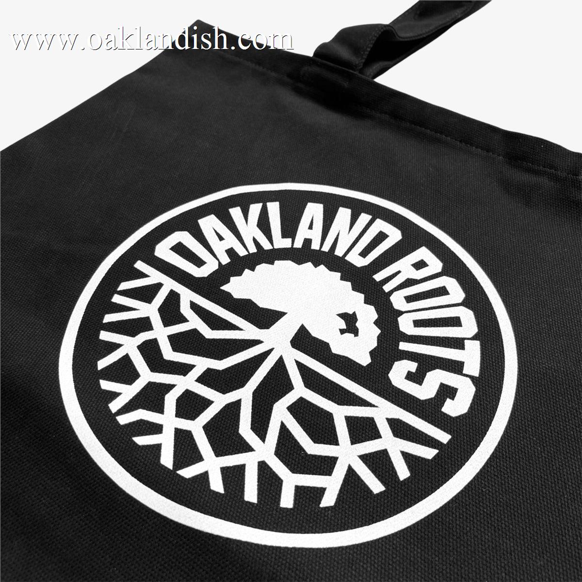 The Oakland Roots SC Tote from Oakland Roots SC showcases a striking white circular logo against its black fabric. The design features bold text spelling 'Oakland Roots' encircling a tree with branching roots, capturing the essence of an Oaklandish vibe. It also includes a single black strap for easy carrying.