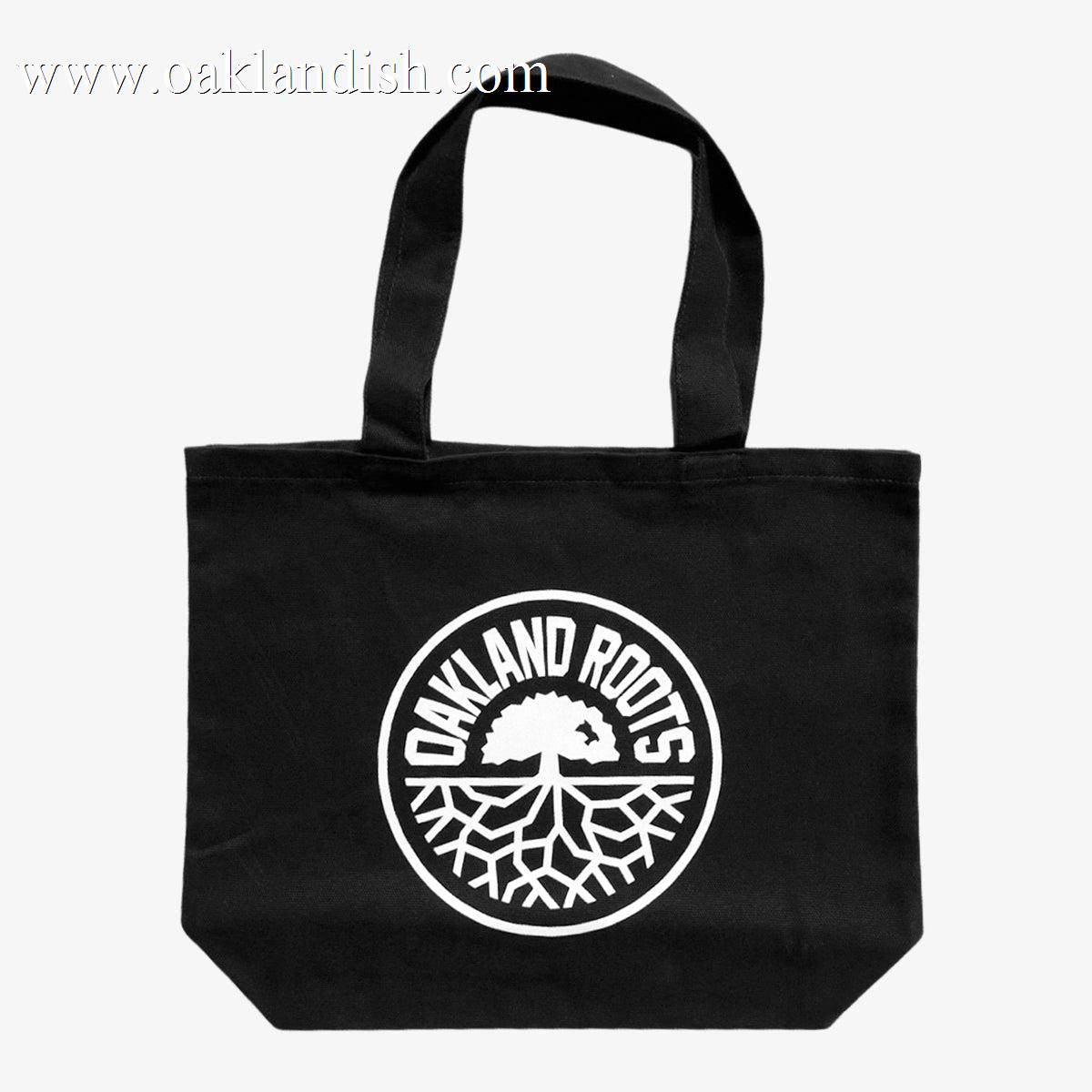 Introducing the Oakland Roots SC Tote by Oakland Roots SC: This black tote bag showcases a circular white emblem at its center, featuring a stylized image of a tree with spreading roots. Encircling the design are the bold, uppercase words "OAKLAND ROOTS." Ideal for fans of the local sports club, it includes two black handles for easy carrying.