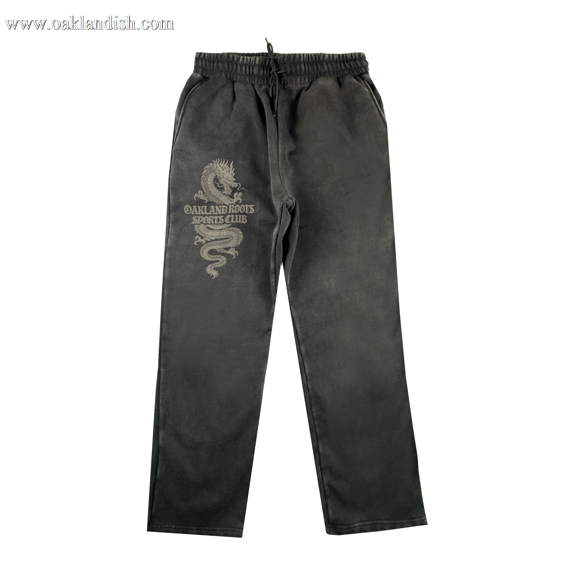 The Tatsu Pants by Oakland Roots SC are black organic cotton sweatpants with an elastic waistband, drawstring, side pockets, a relaxed straight-leg fit, and a light dragon graphic plus "OAKLAND ROOTS SPORTS CLUB" on the left thigh.