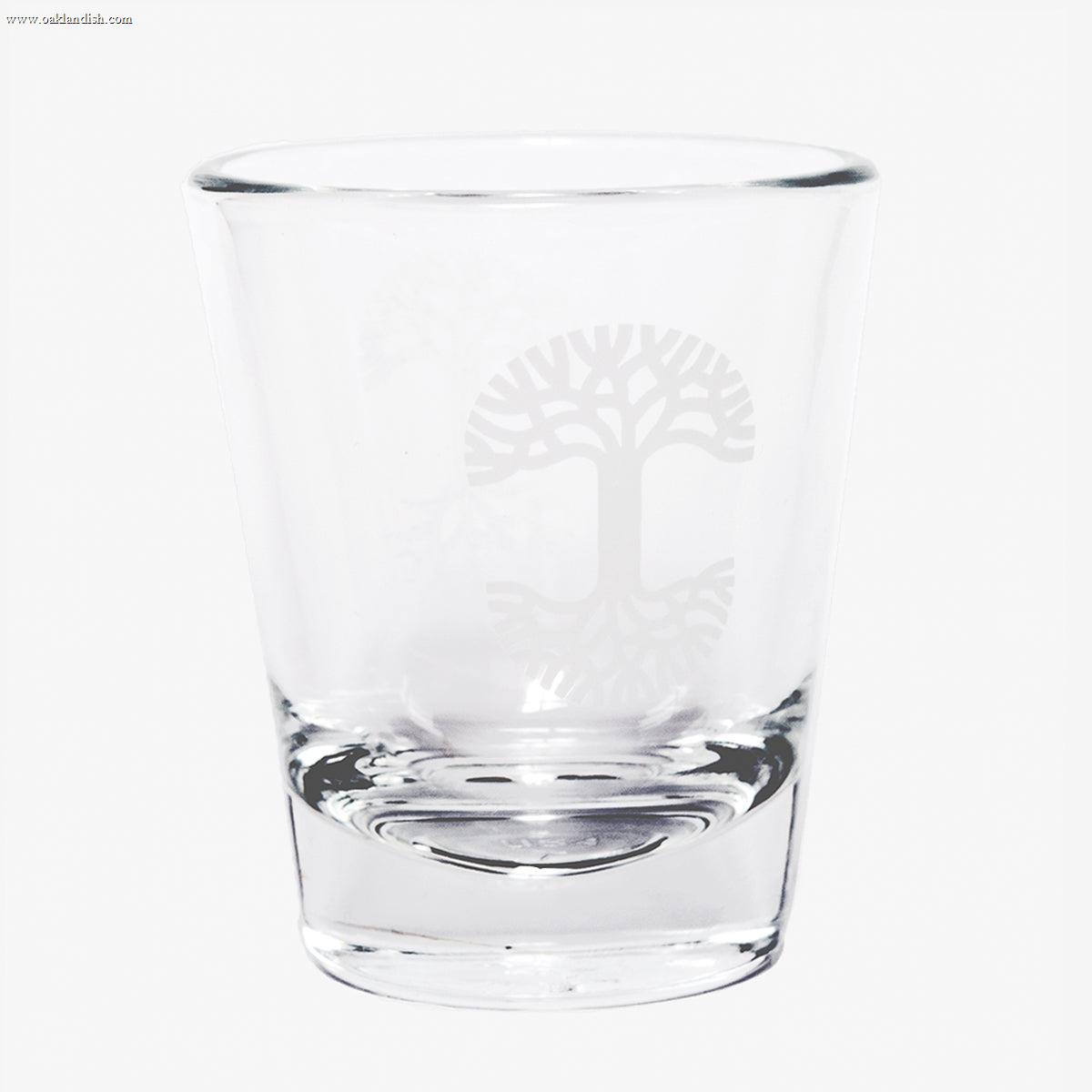 The Oaklandish Trees Shot Glass by Oaklandish is a clear glass tumbler with a thick, solid base, showcasing an elegant logo etching of a stylized tree with sprawling branches and roots, symbolizing the tree of life. This exquisite shot glass is empty and set against a plain white background.