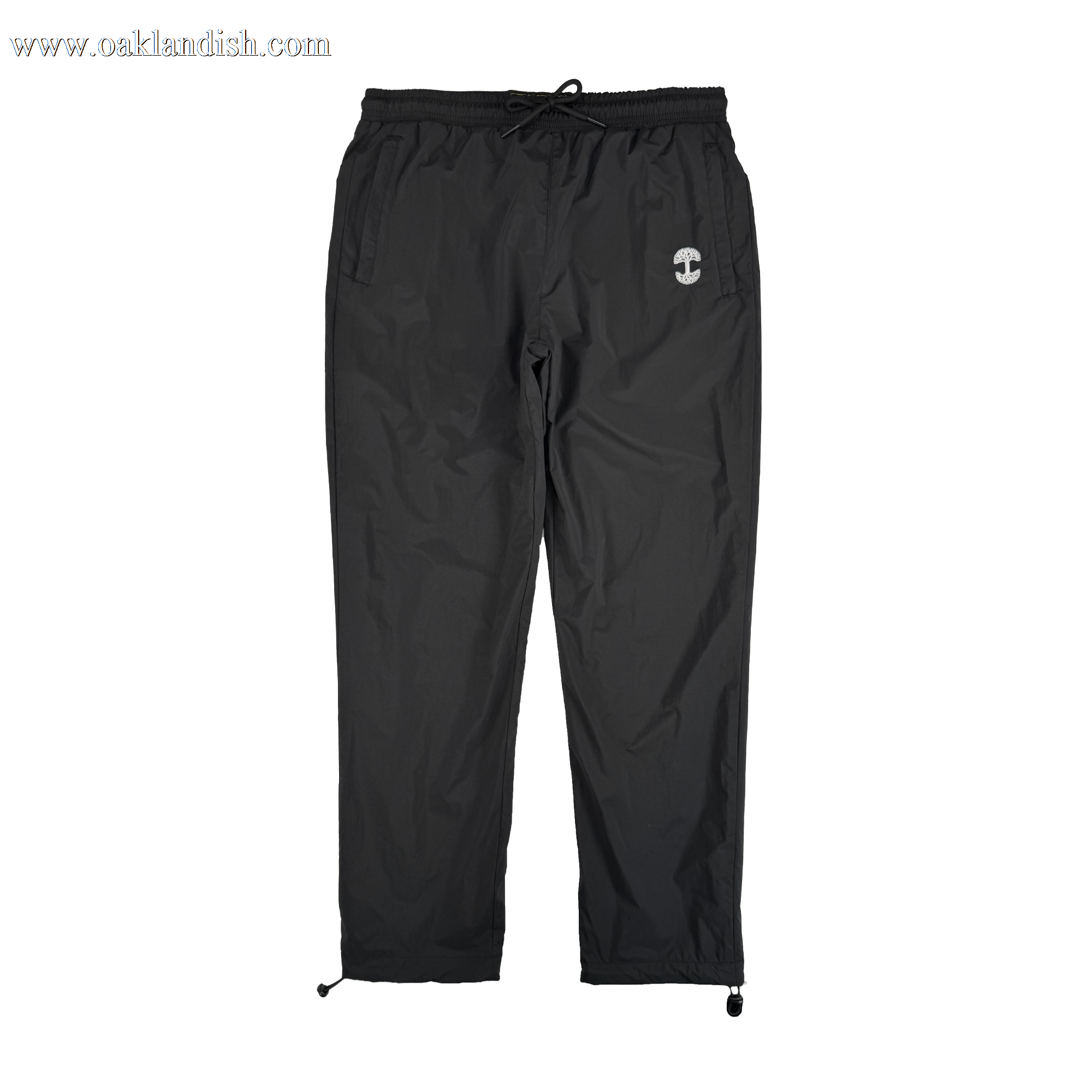 The Oaklandish Susurrus Track Bottom is a pair of black nylon pants featuring an adjustable waistband, drawstring cuffs, two side pockets, and a small white circular logo on the left thigh. Set against a plain white background.