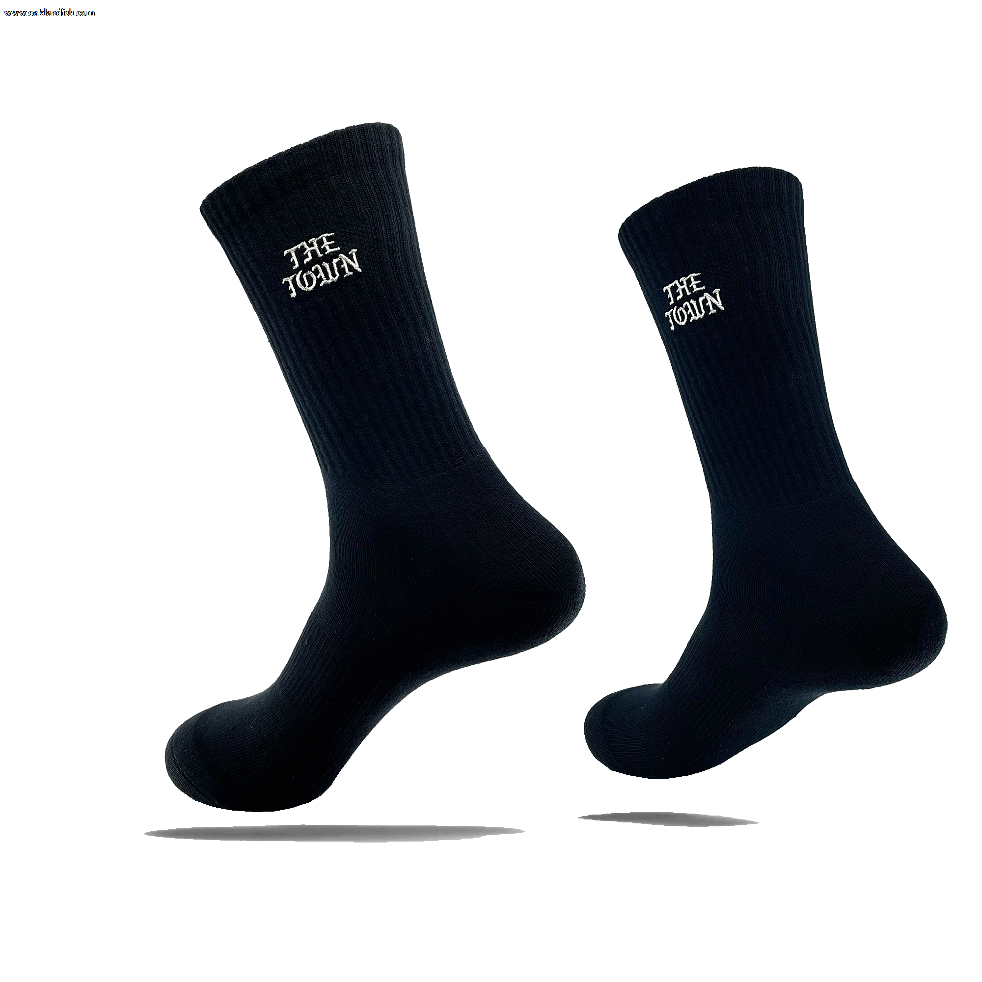 Image of two black crew socks, each showcasing the white text "THE TOWEL" near the top. These stylish crew socks, named The Town Old English Sock by Oaklandish, are one size fits most and appear to be floating against a transparent background, casting a shadow beneath each sock. The material features a ribbed texture at the top.