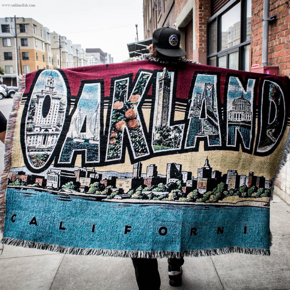 A person wearing a black cap and dark clothing walks down a sidewalk, carrying the large "Greetings From Oakland Throw" by Oaklandish. The tapestry, crafted from recycled fibers, prominently features the word "Oakland" in bold letters and depicts various city landmarks in Oakland, California, set against a vibrant background of blue, tan, and burgundy.