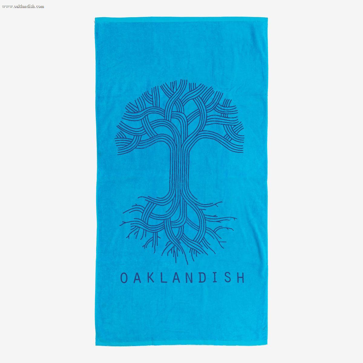 The Classic Logo Beach Towel from Oaklandish features a bright blue, oversized plush design with a stylized drawing of a tree with intertwining branches and roots created with bold black lines. Below the tree, the text "OAKLANDISH" is printed in black capital letters. Crafted from cotton terry velour, this rectangular towel also boasts a plain background.