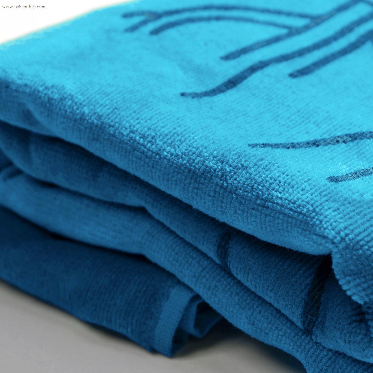Close-up of a stack of folded, bright blue Classic Logo Beach Towels by Oaklandish, featuring a decorative stitched pattern. The texture of these oversized plush cotton terry velour towels appears soft and lush. The neatly arranged towels rest on a smooth, light-colored surface.