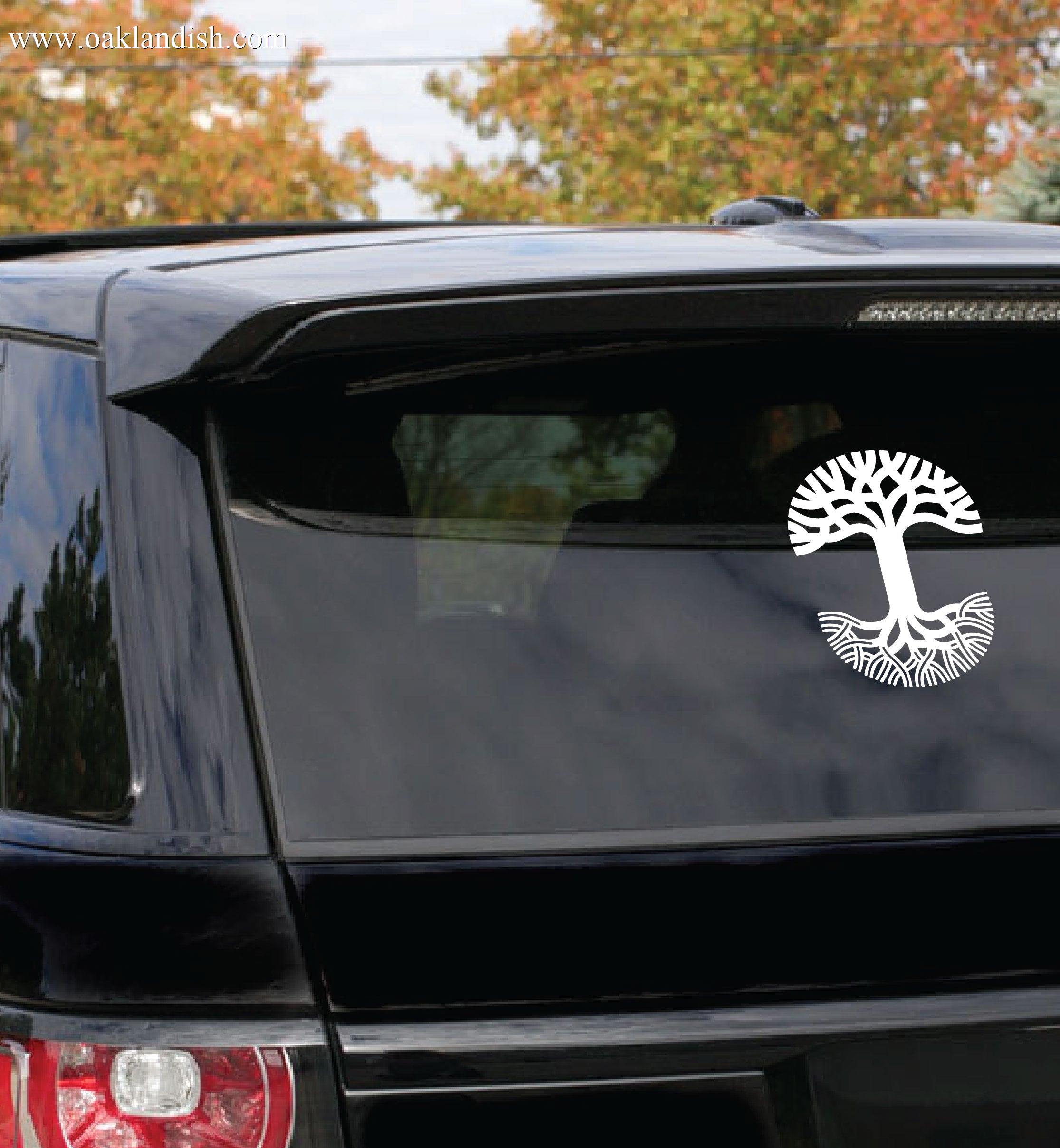 An outdoor setting in Oakland features a black SUV parked among trees adorned with autumn foliage. On the rear window of the SUV is an Oaklandish Decal 8x10 by Oaklandish, showcasing a white tree design with extensive roots forming a circular pattern and branches spreading upwards to cover the top half.