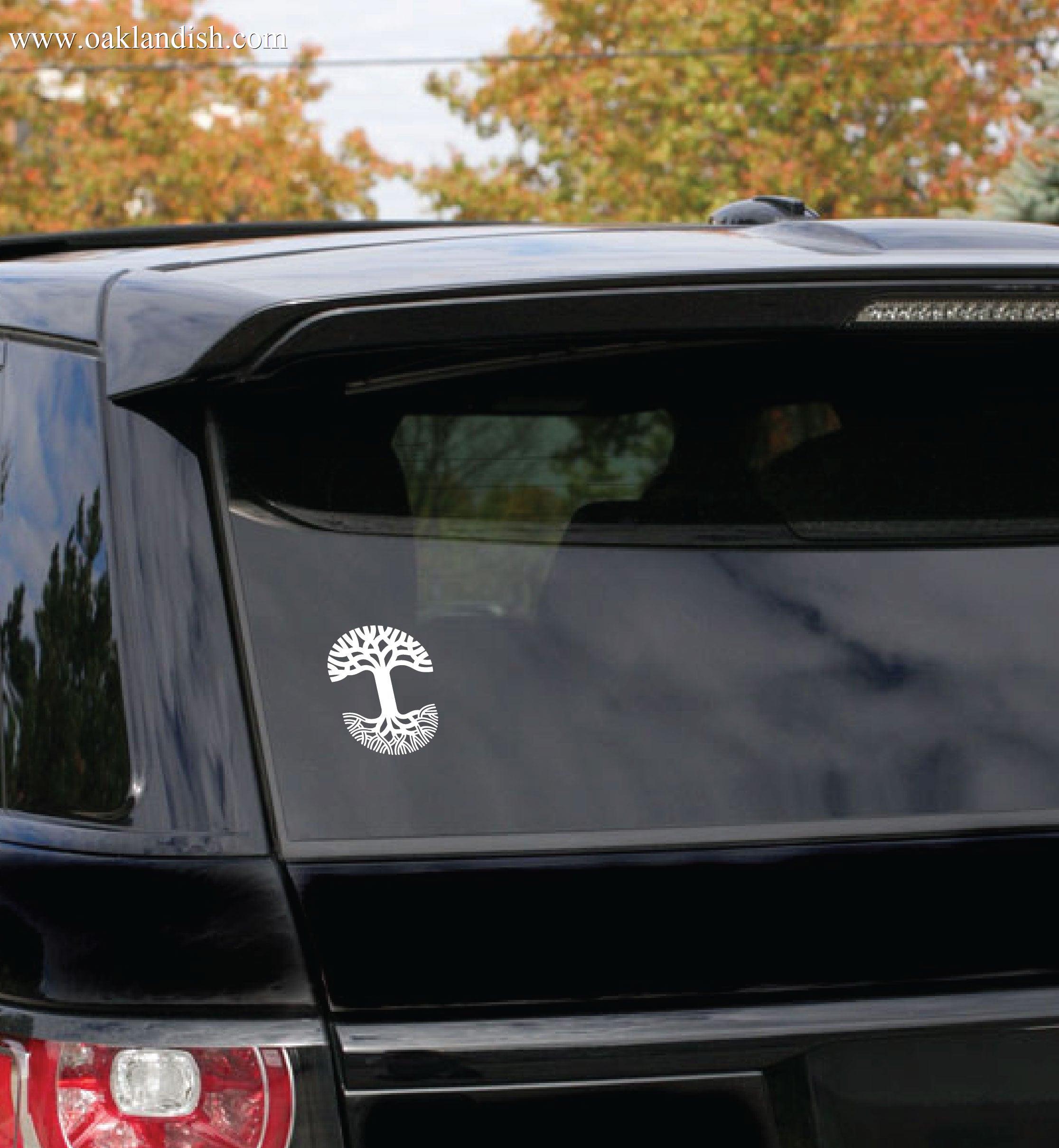 A durable black SUV with an Oaklandish Decal 5x7 white tree sticker on the rear window is parked outdoors. In the background, trees with autumn foliage are visible under a partly cloudy sky.