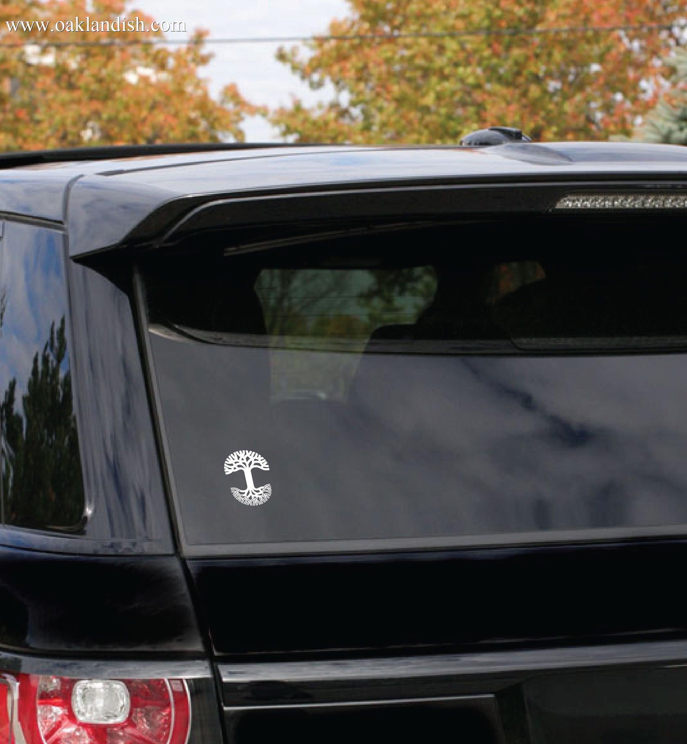 A black SUV is parked with its rear window visible, adorned with a small white Oaklandish Decal 3X4.5 depicting the Tree of Gondor. The trees in the background suggest an autumn setting, with their leaves showing various shades of orange and green. The vehicle is partially in shade, reflecting clear autumn light on its durable outdoors finish.
